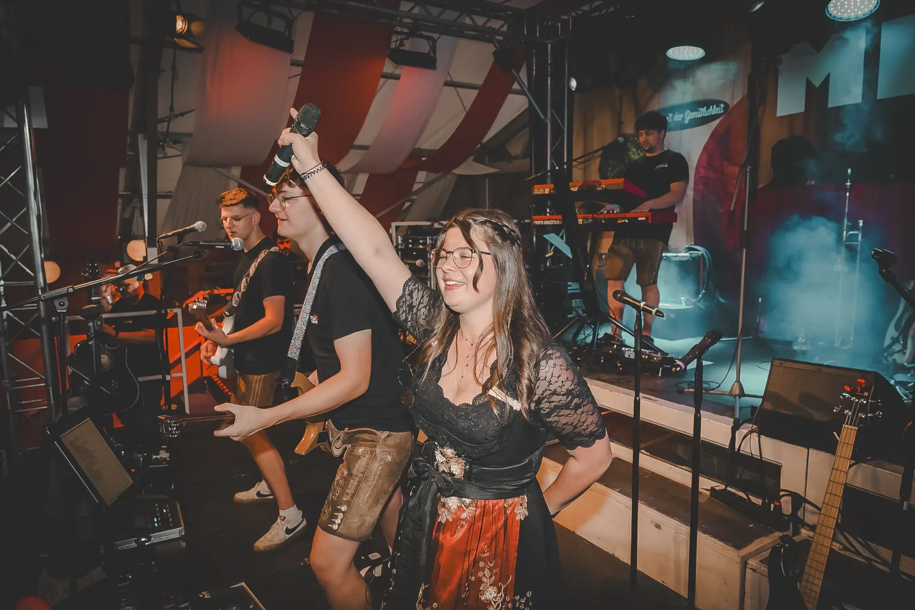 Liveband auf einer Festzeltbühne: Eine Sängerin im Dirndl hebt lächelnd ein Mikrofon in die Luft, während zwei Gitarristen daneben spielen und ein Keyboarder im Hintergrund zu sehen ist.