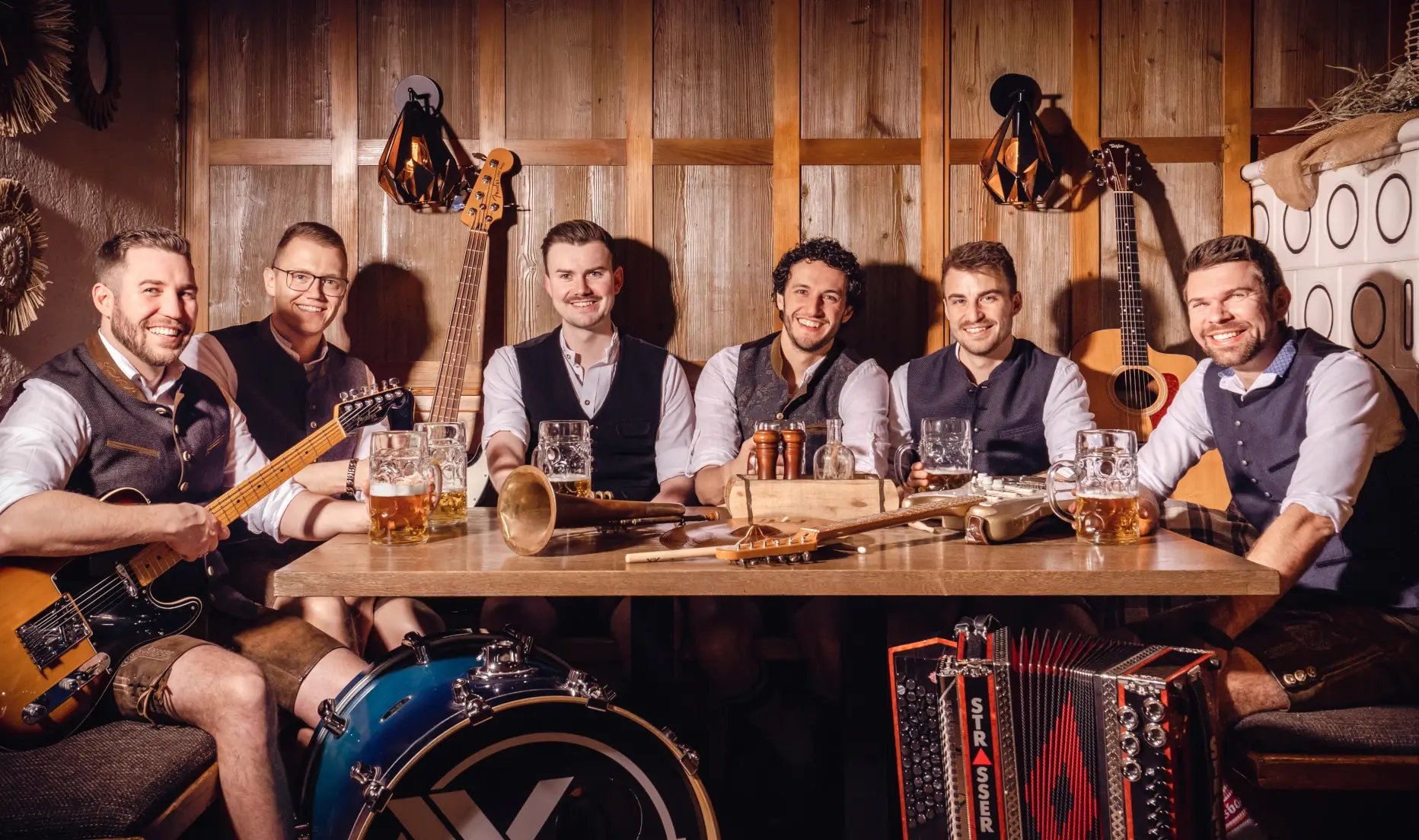 Bayrische Partyband X'Ploushn mit sechs Musikern in Tracht an einem Holztisch mit Instrumenten in rustikalem Ambiente