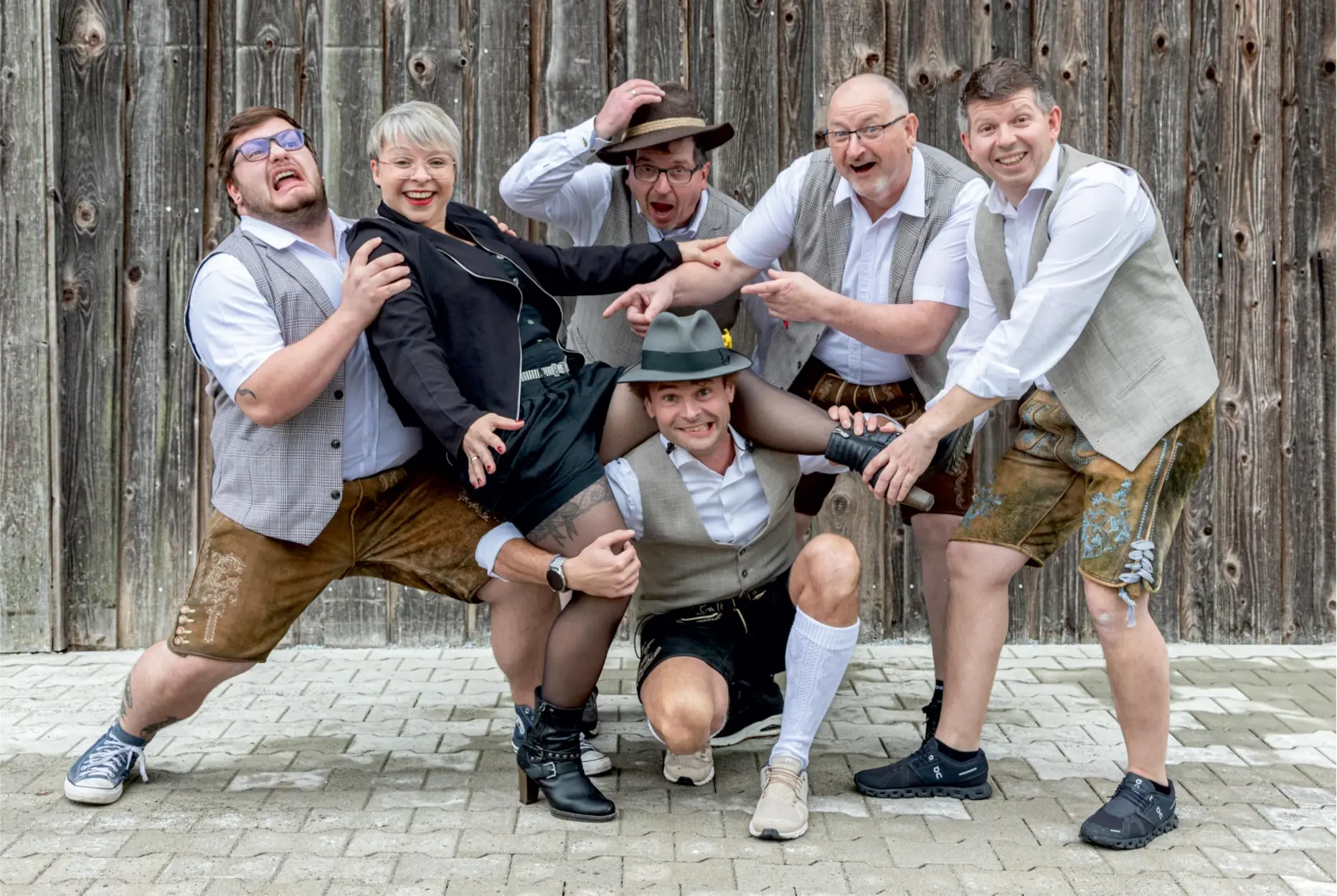 De Zwiadn – die bayrische Partyband aus Furth im Wald, sechs Musiker in Tracht und moderner Kleidung posieren ausgelassen vor einer Holzwand.