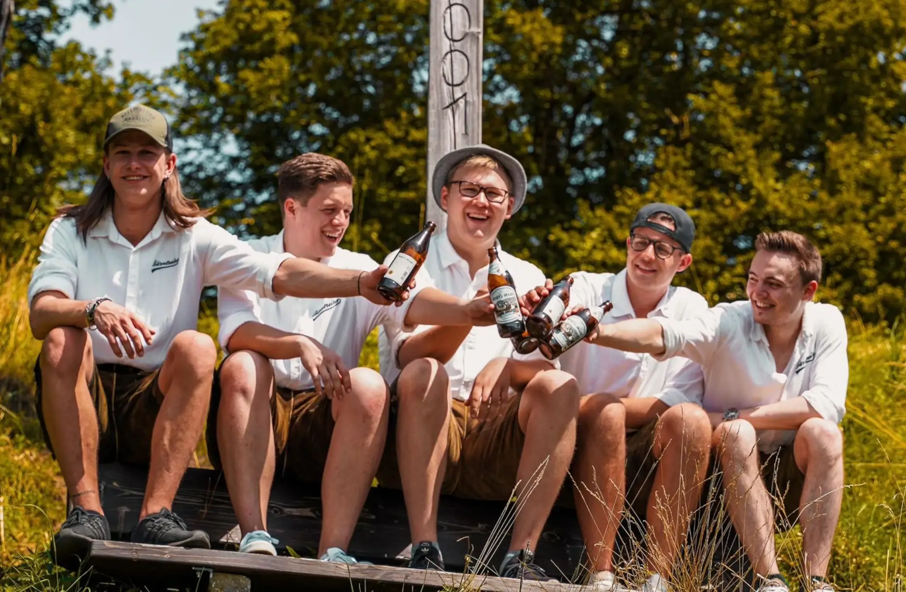 Bärntreiber – die bayrische Partyband aus Velburg, fünf Musiker in Tracht stoßen mit Bierflaschen an und genießen die Sonne auf einer Wiese.