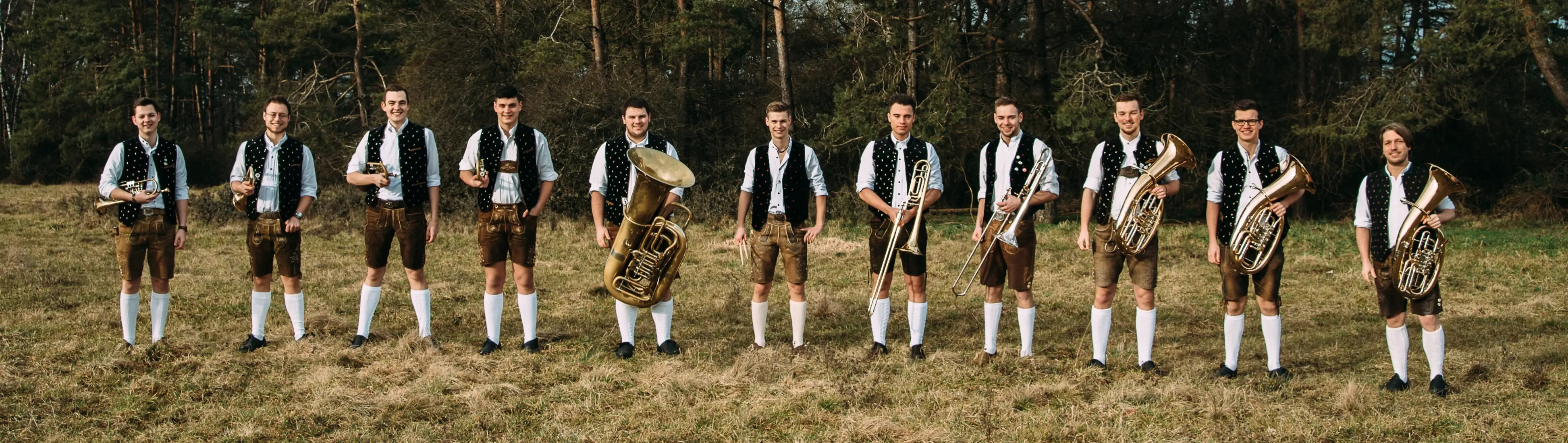 Die Musiker der Blaskapelle Dezent Böhmisch gehen aufgereiht nebeneinander über eine Wiese mit ihren Instrumenten in den Händen, dahinter ein Wald.