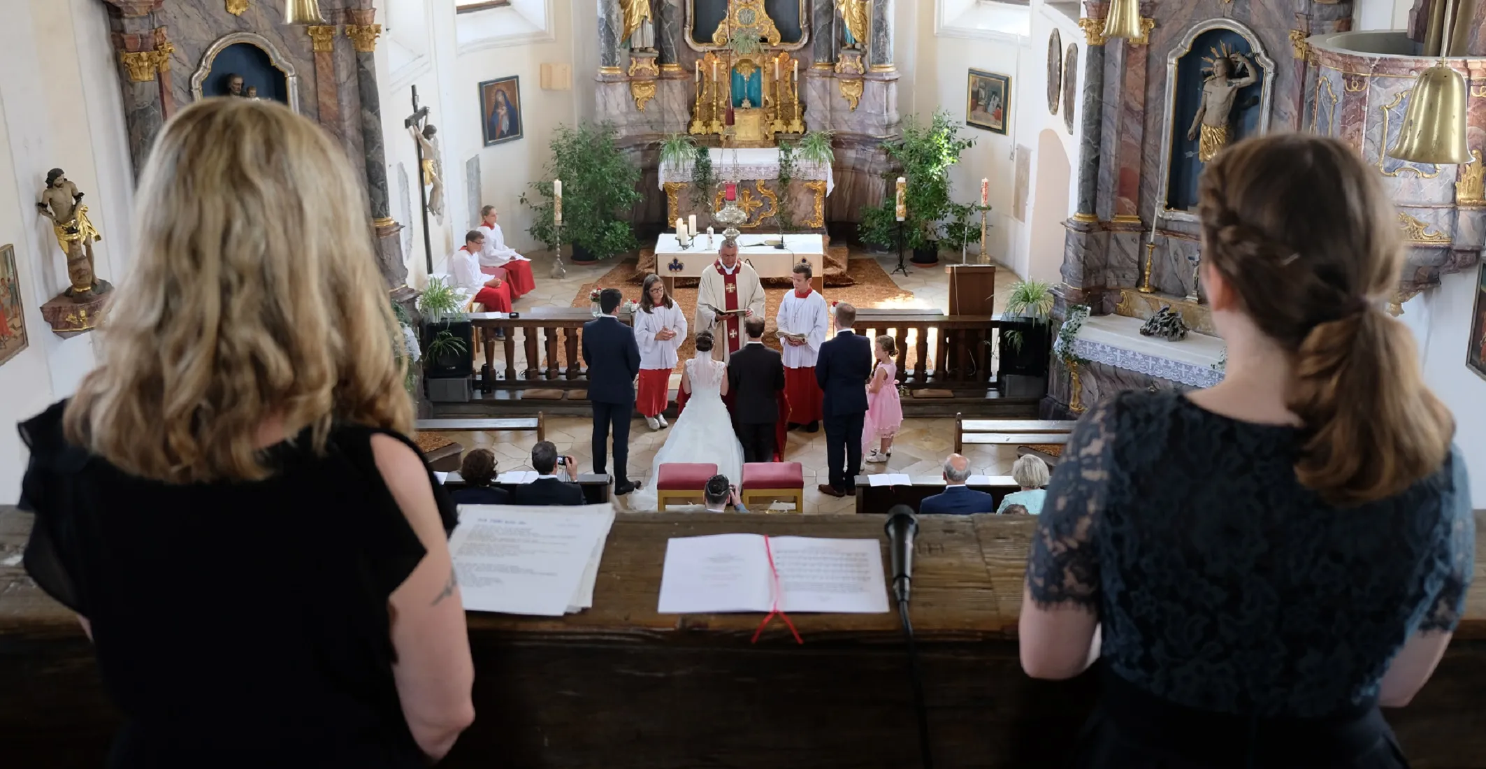 Unscharf sieht man Micro, Noten und die beiden Sängerinnen der Band Freunde des Brautpaares auf der Empore von der herab man ein Brautpaar mit Pfarrer bei einer Hochzeit erkennen kann.