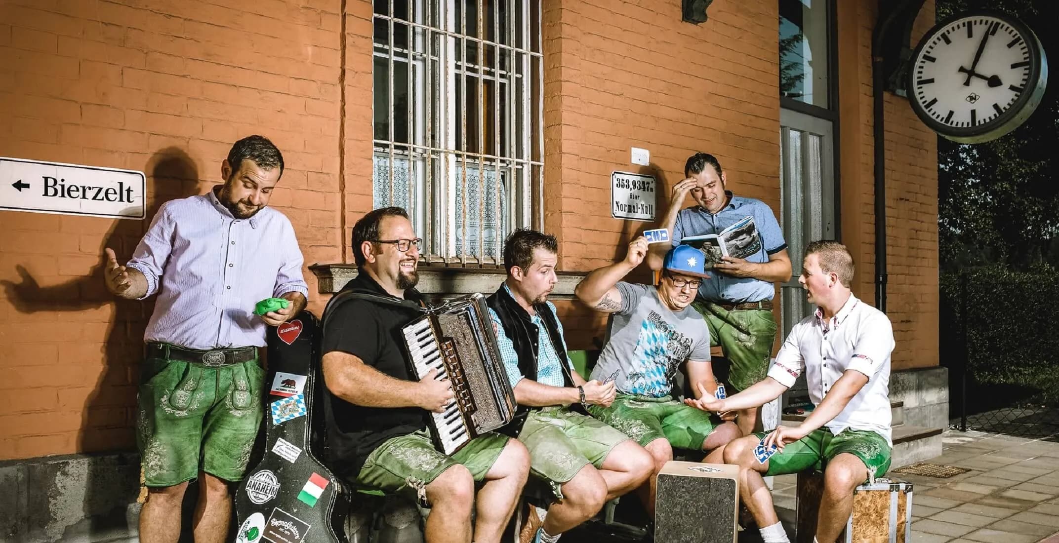 Froschhaxn Express – die bayrische Partyband aus Schmidmühlen, sechs Musiker in grüner Lederhose beim Kartenspielen und Musizieren vor einem Bahnhof mit Bierzelt-Schild – perfekt für Festzelt, Volksfest oder Gründungsfest.