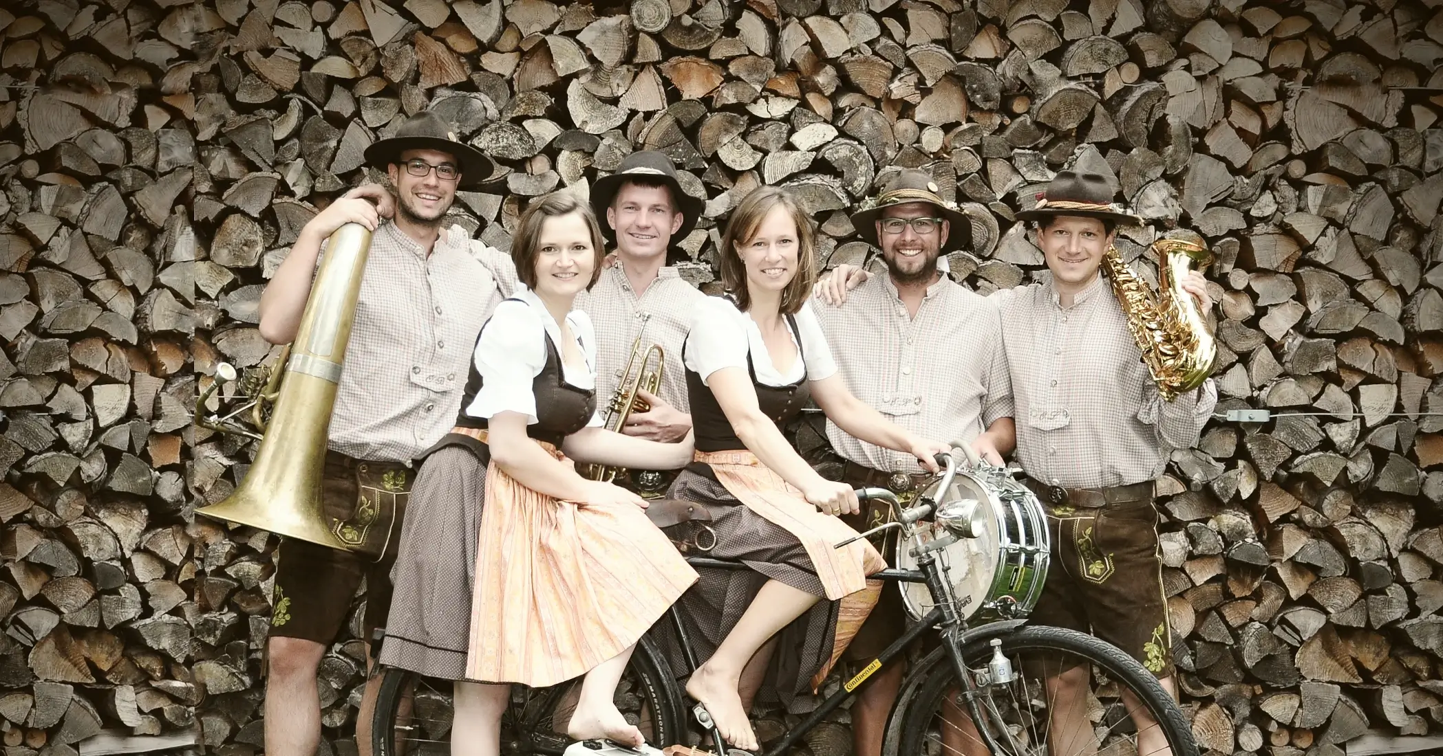 Let’s Fetz – die Tanz- und Showband aus Adelshausen in traditioneller Tracht mit Tuba, Trompete, Saxophon und Trommel, fröhliches Bandfoto vor Holzstapeln mit altem Fahrrad.