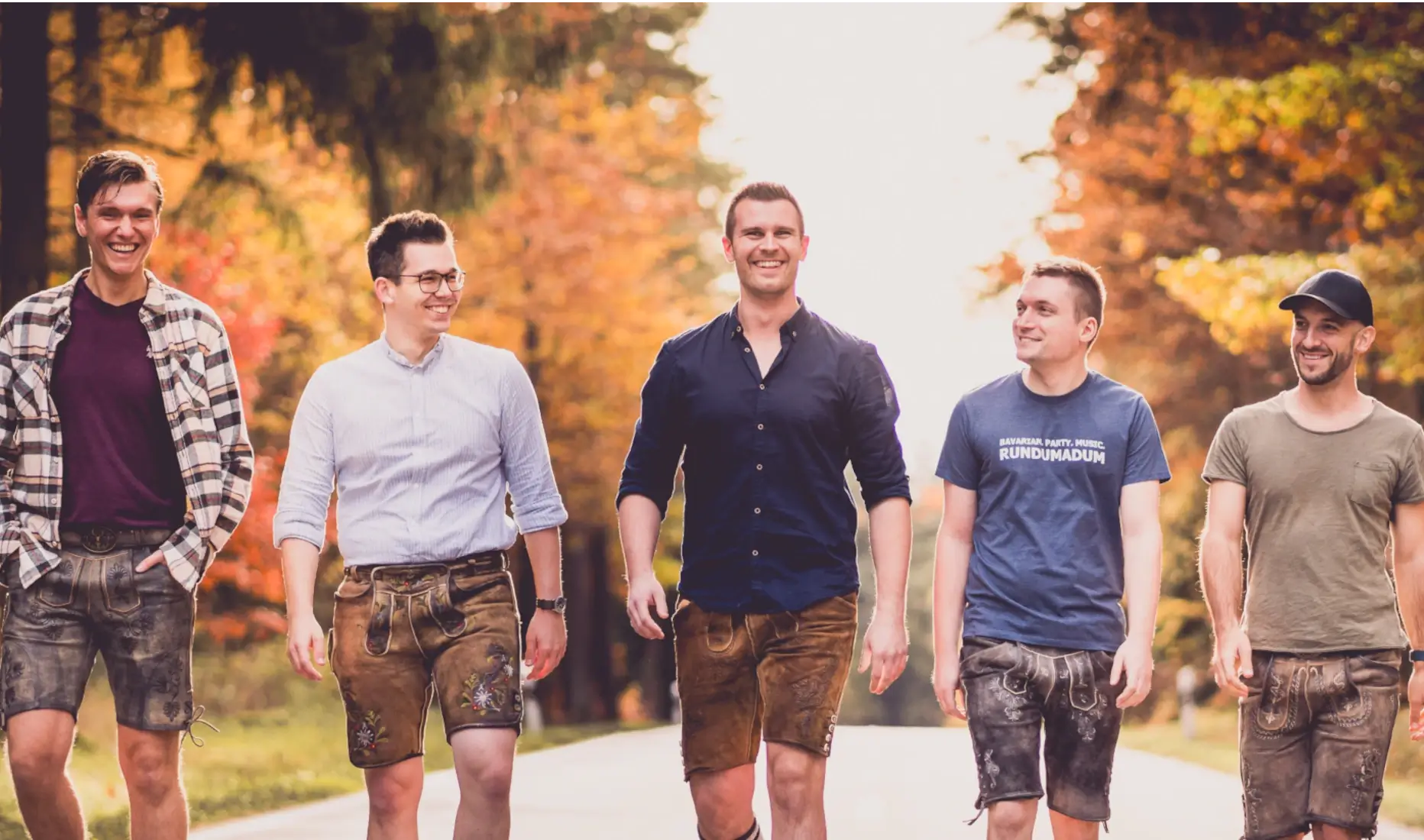 Rundumadum – die bayrische Partyband aus Fensterbach, fünf Musiker in Lederhosen und lässiger Kleidung spazieren fröhlich eine herbstliche Landstraße entlang.