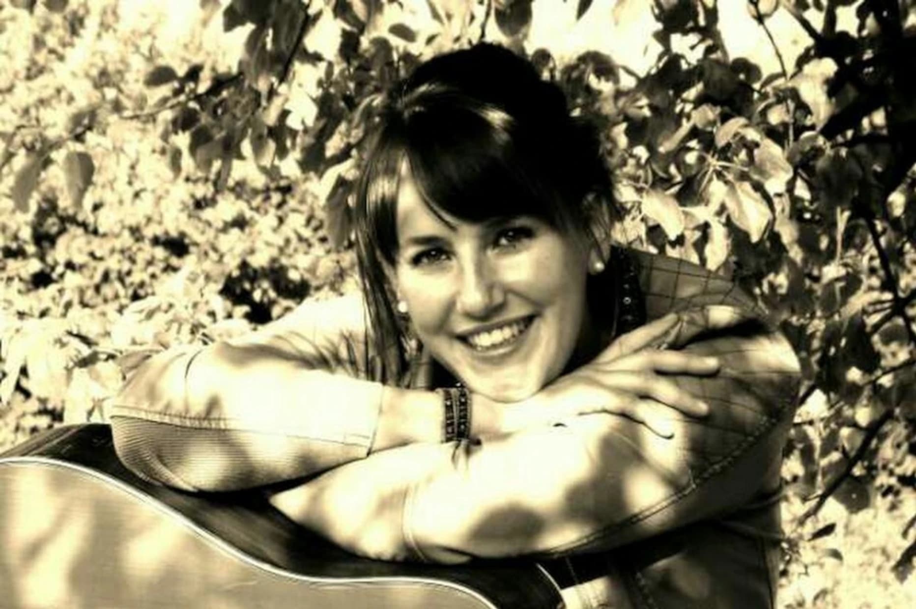 Die Musikern Sabrina Robold nachdenklich sitzend mit der Gitarre auf den Oberschenkeln vor einem Baum.