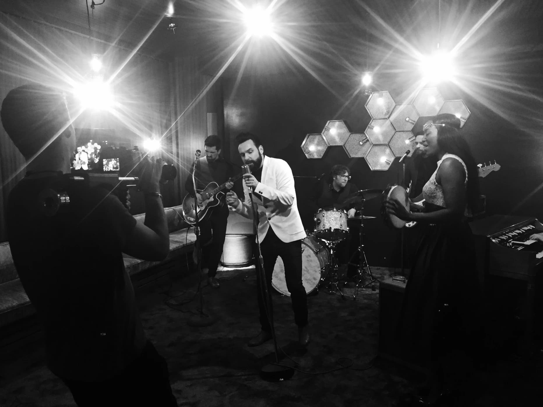San2 mit Band bei einer Live-Session: Sänger San2 im weißen Jacket am Mikrofon, begleitet von Gitarre, Schlagzeug, Bass und einer Sängerin mit Tamburin, während ein Kameramann die Performance filmt. Schwarz-weiß-Aufnahme mit starker Bühnenbeleuchtung.