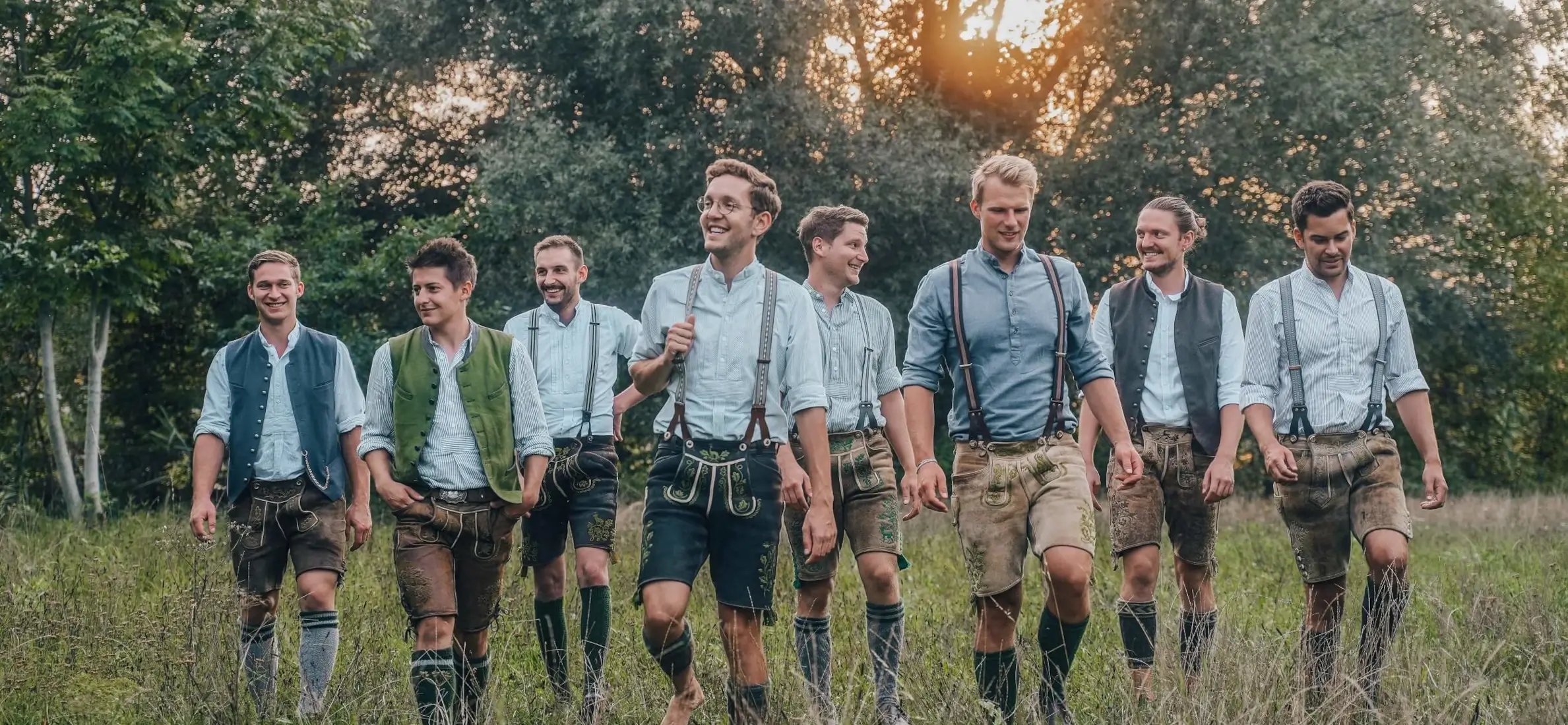 Saustoimusi – die bayrische Partyband aus Hepberg, neun junge Musiker in traditioneller Tracht spazieren lachend durch eine Wiese bei Sonnenuntergang – ideal für Festzelt, Volksfest oder Gründungsfest.