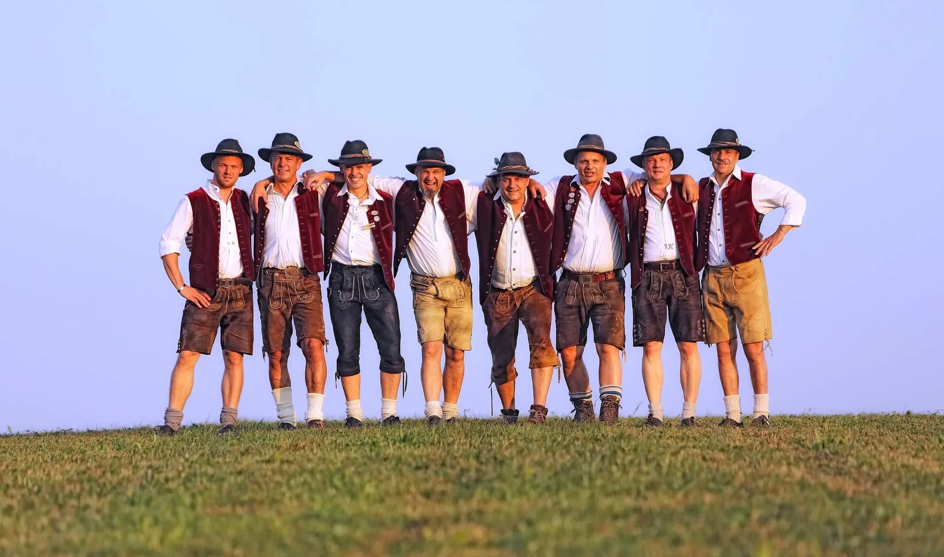SIMMISAMMA – die Oktoberfestband aus Flintsbach, acht Musiker in traditioneller Tracht mit Lederhosen und Westen posieren im Sonnenuntergang auf einer Wiese – ideal für Volksfeste, Oktoberfest und FF-Jubiläen.