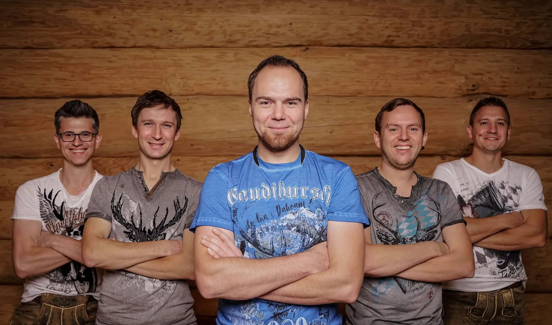 Spitz af Knopf – die bayrische Partyband aus Parsberg, fünf Musiker in Tracht-inspirierten Shirts posieren vor einer Holzwand mit Bandlogo.